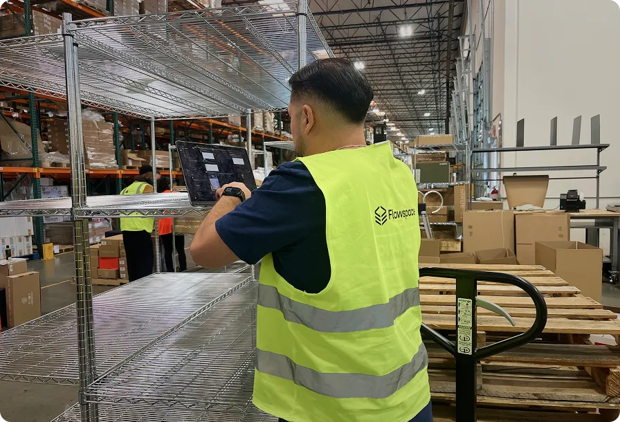 Warehouse worker in a yellow safety vest using a tablet while inspecting metal shelves in a large storage facility.
