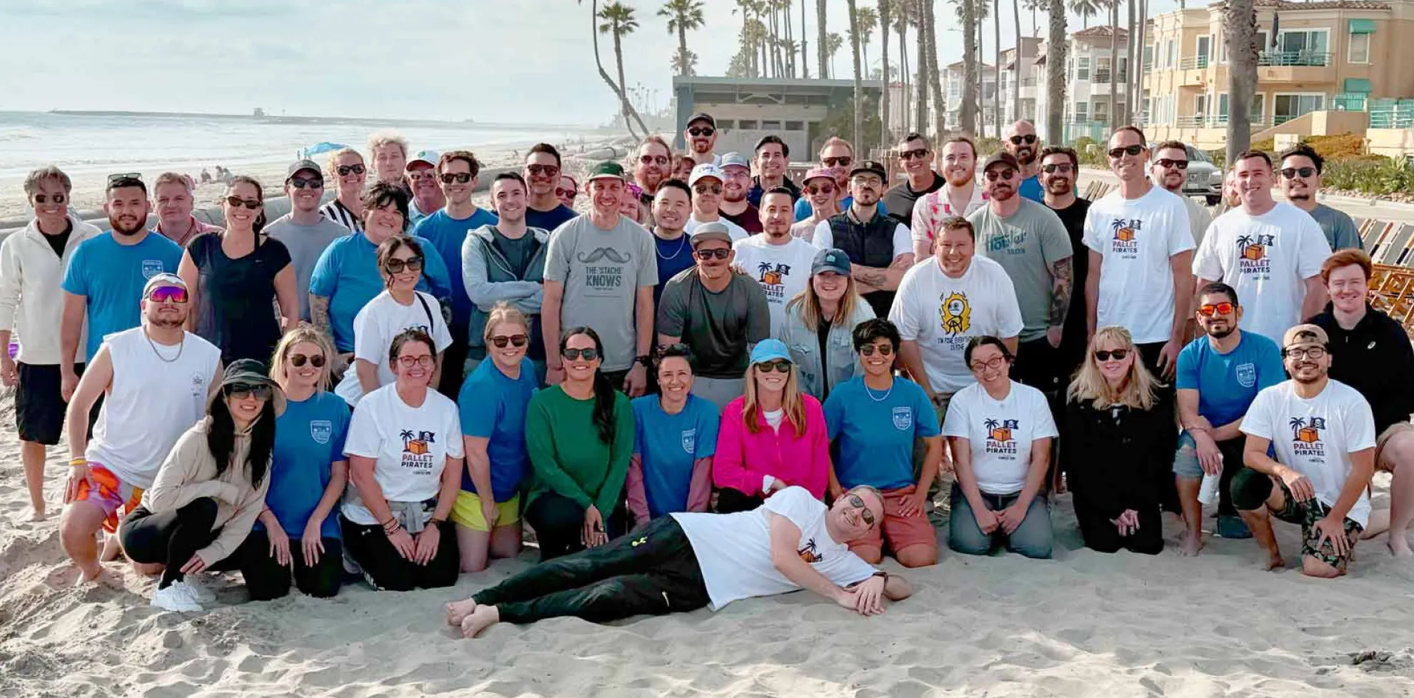 Large group of people posing together on a beach with palm trees and buildings in the background.