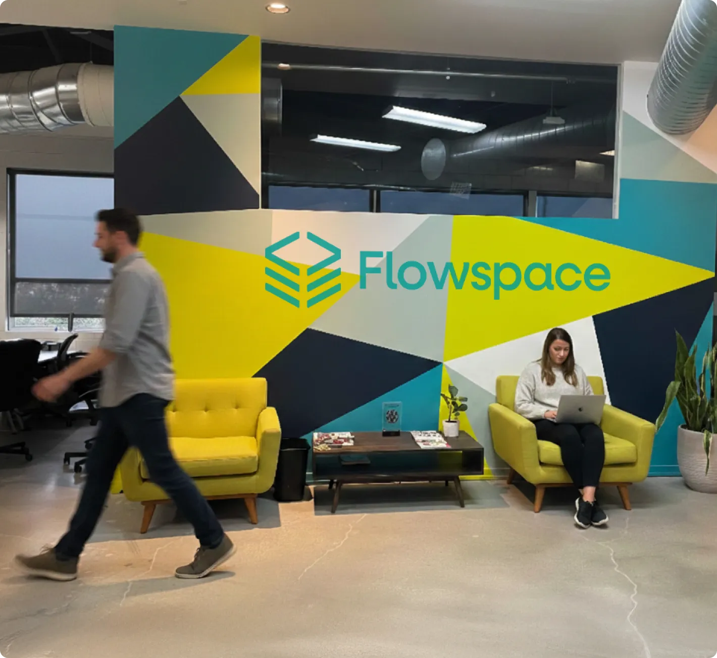 Modern office reception area with colorful geometric wall art and 'Flowspace' logo, featuring two yellow armchairs, one occupied by a woman using a laptop and a man walking past.