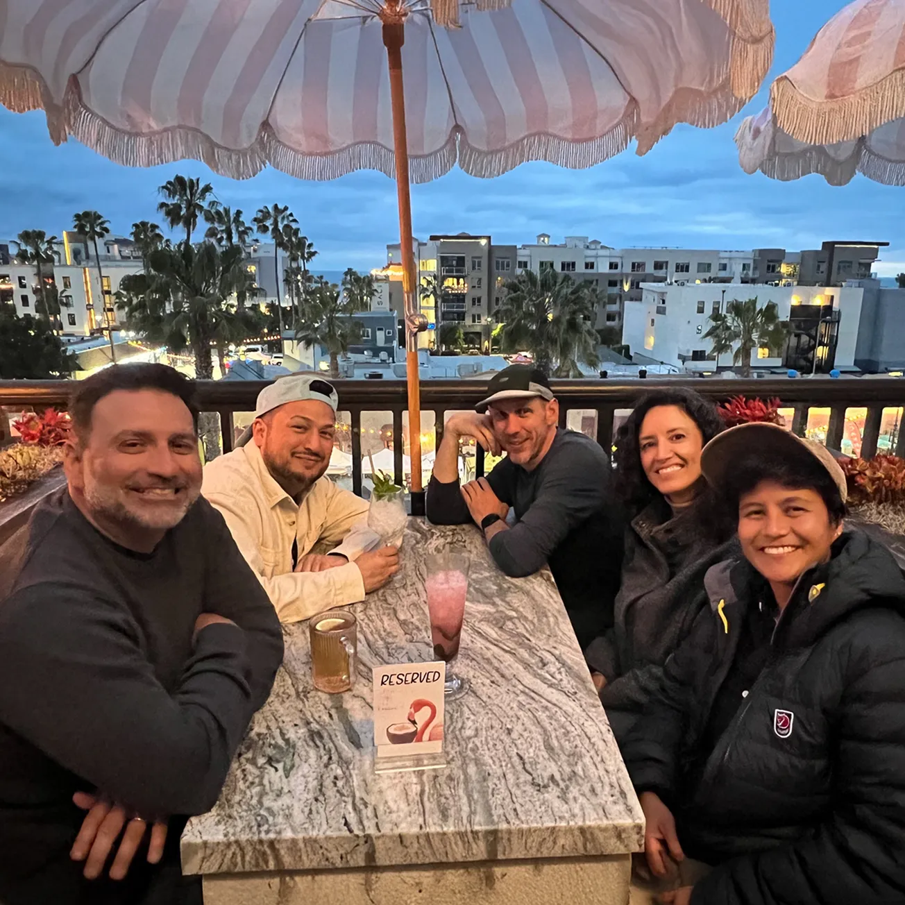 Five people sitting around a marble table under an outdoor umbrella at dusk with drinks, smiling and posing for a photo on a balcony overlooking palm trees and buildings.