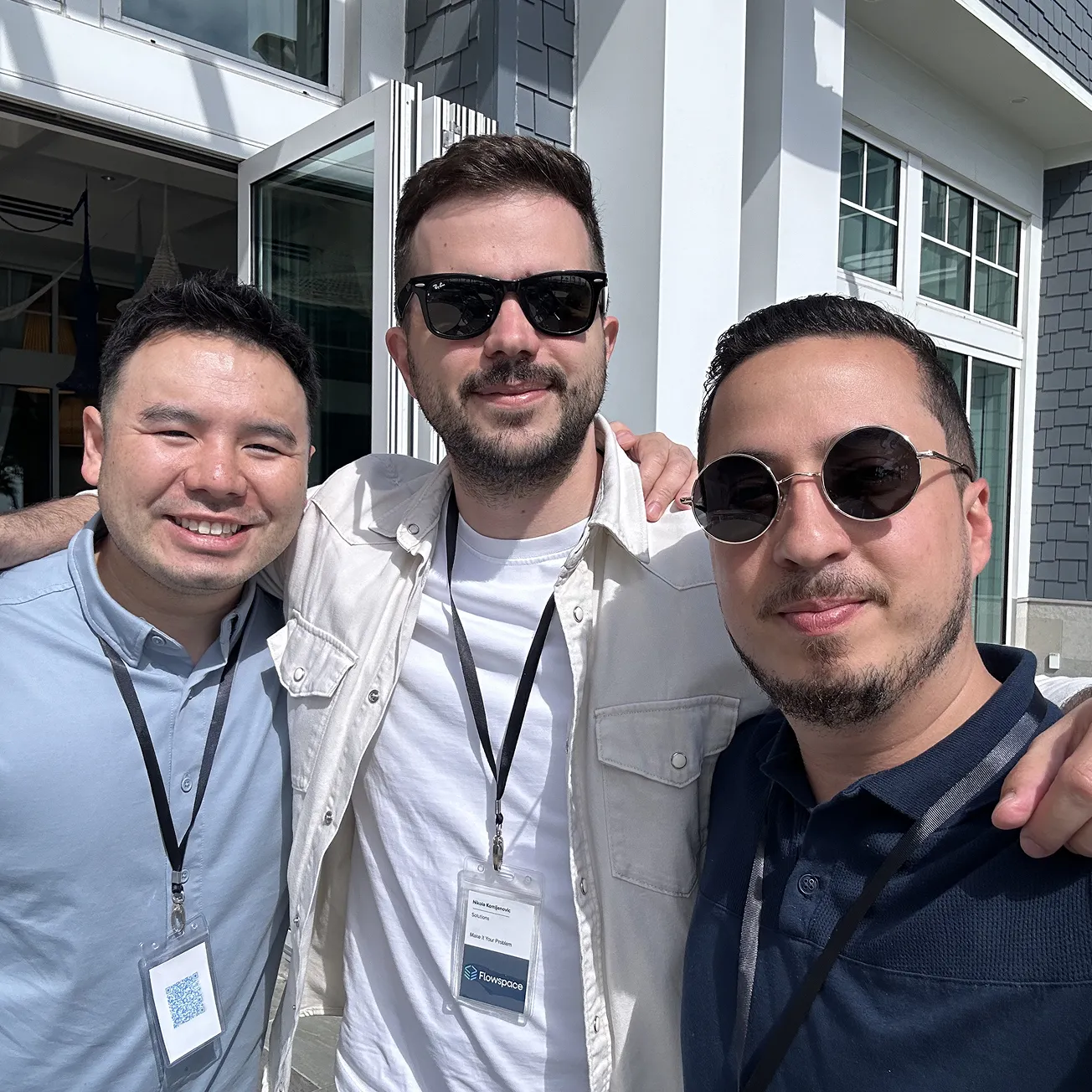 Three men wearing sunglasses and name badges smiling outdoors in front of a building with large windows.