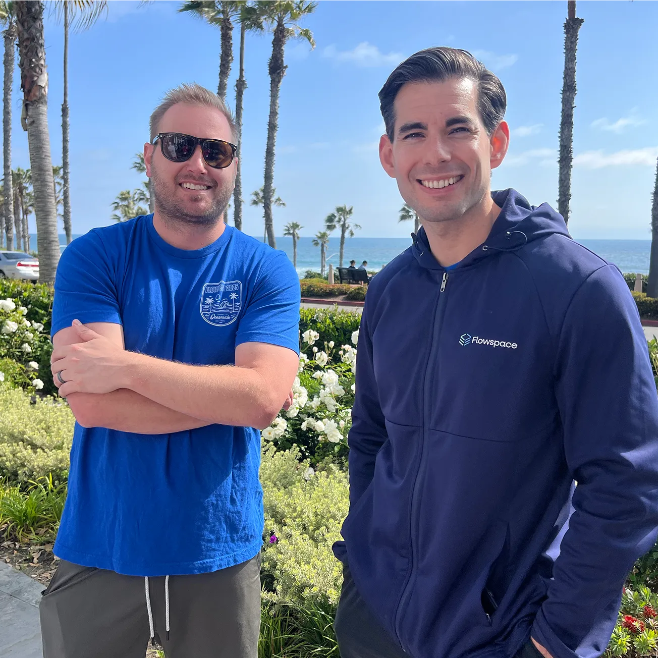 Two men smiling outdoors by a garden with palm trees and ocean in the background, one wearing sunglasses and a blue t-shirt, the other in a navy blue Flowspace jacket.