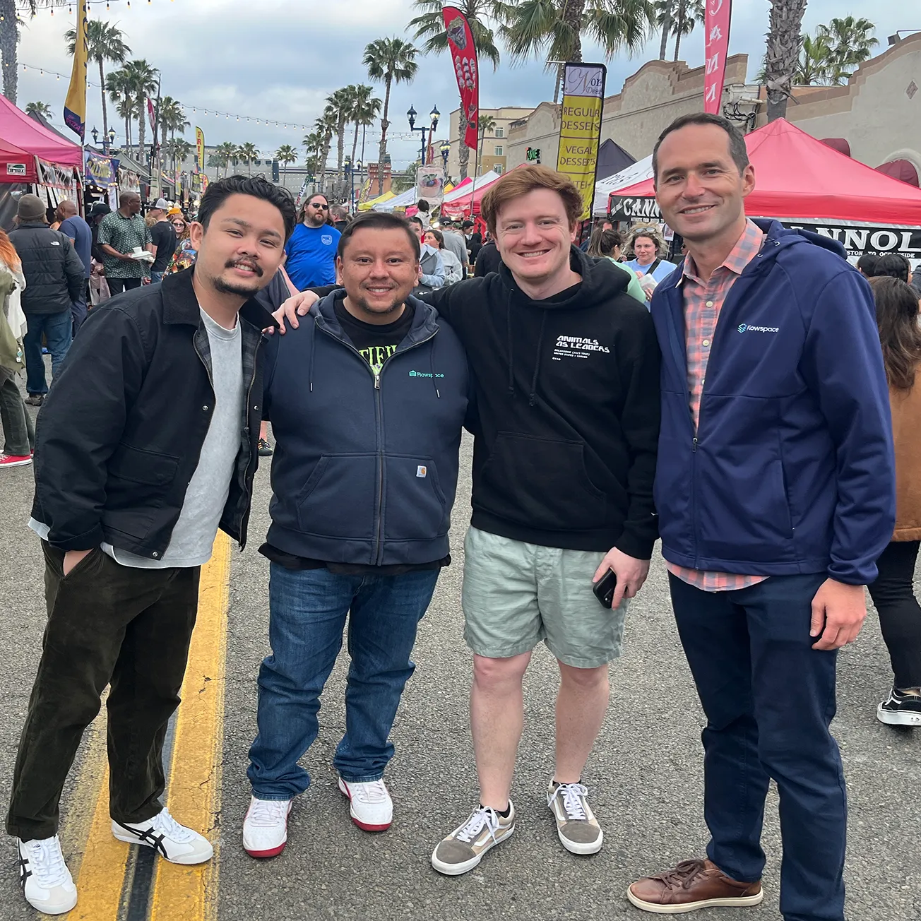 Four men standing close together and smiling at an outdoor market with palm trees, vendor tents, and people in the background.