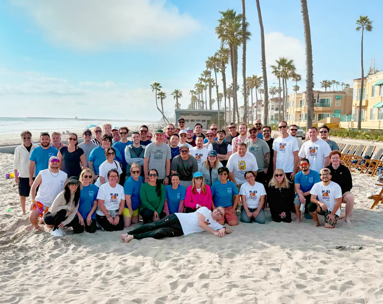 Large group of people posing on a sunny beach with palm trees and ocean in the background.