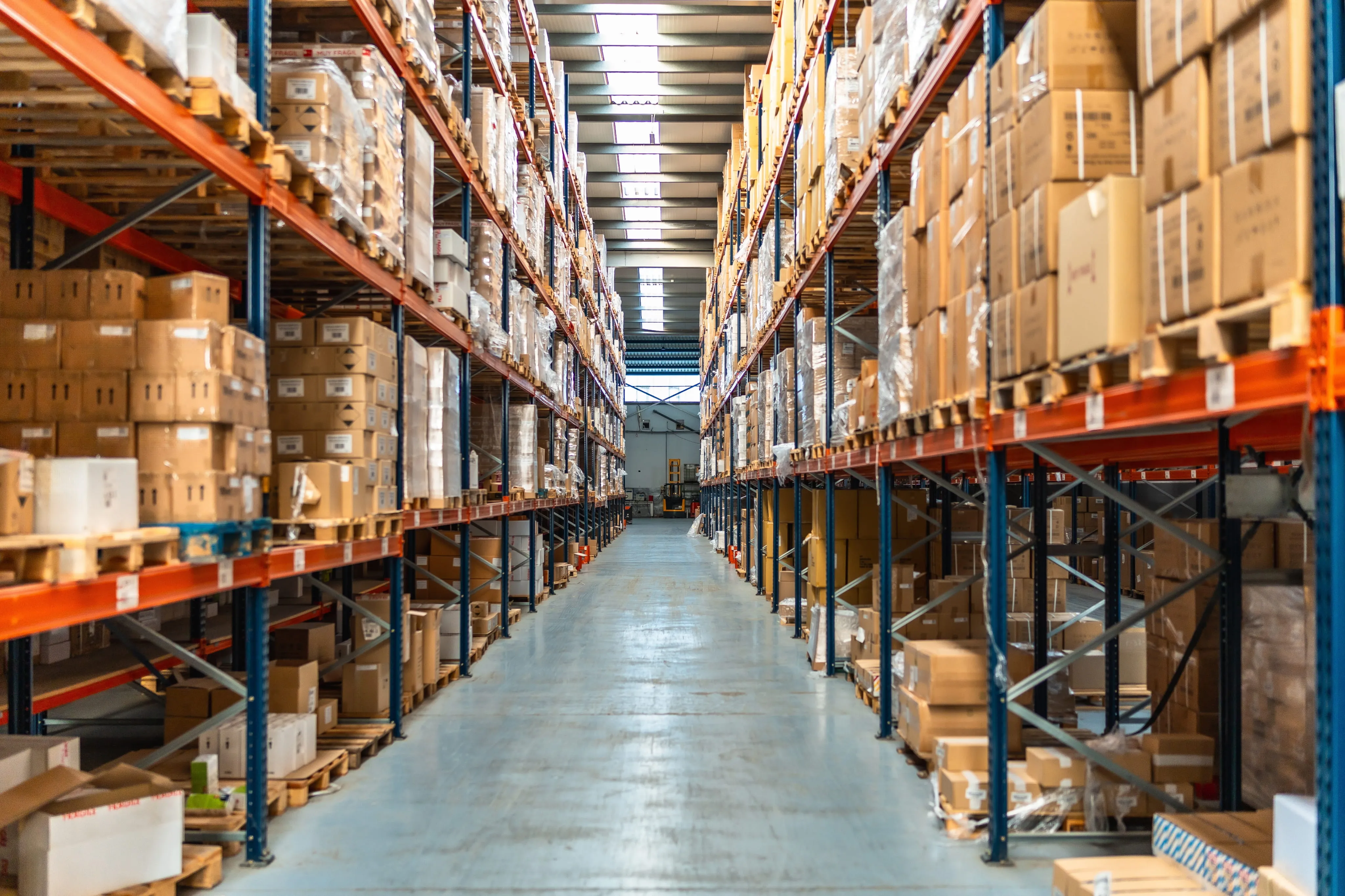 Aisle in a large warehouse lined with tall metal shelves stocked with stacked boxes and pallets.