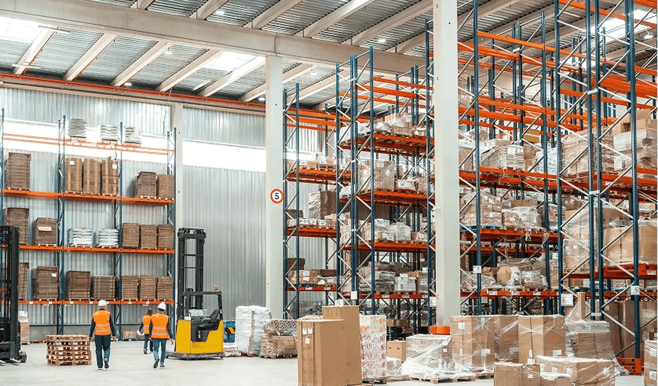 Large warehouse interior with tall orange and blue storage racks filled with boxed inventory and two workers wearing orange safety vests and white helmets walking through.