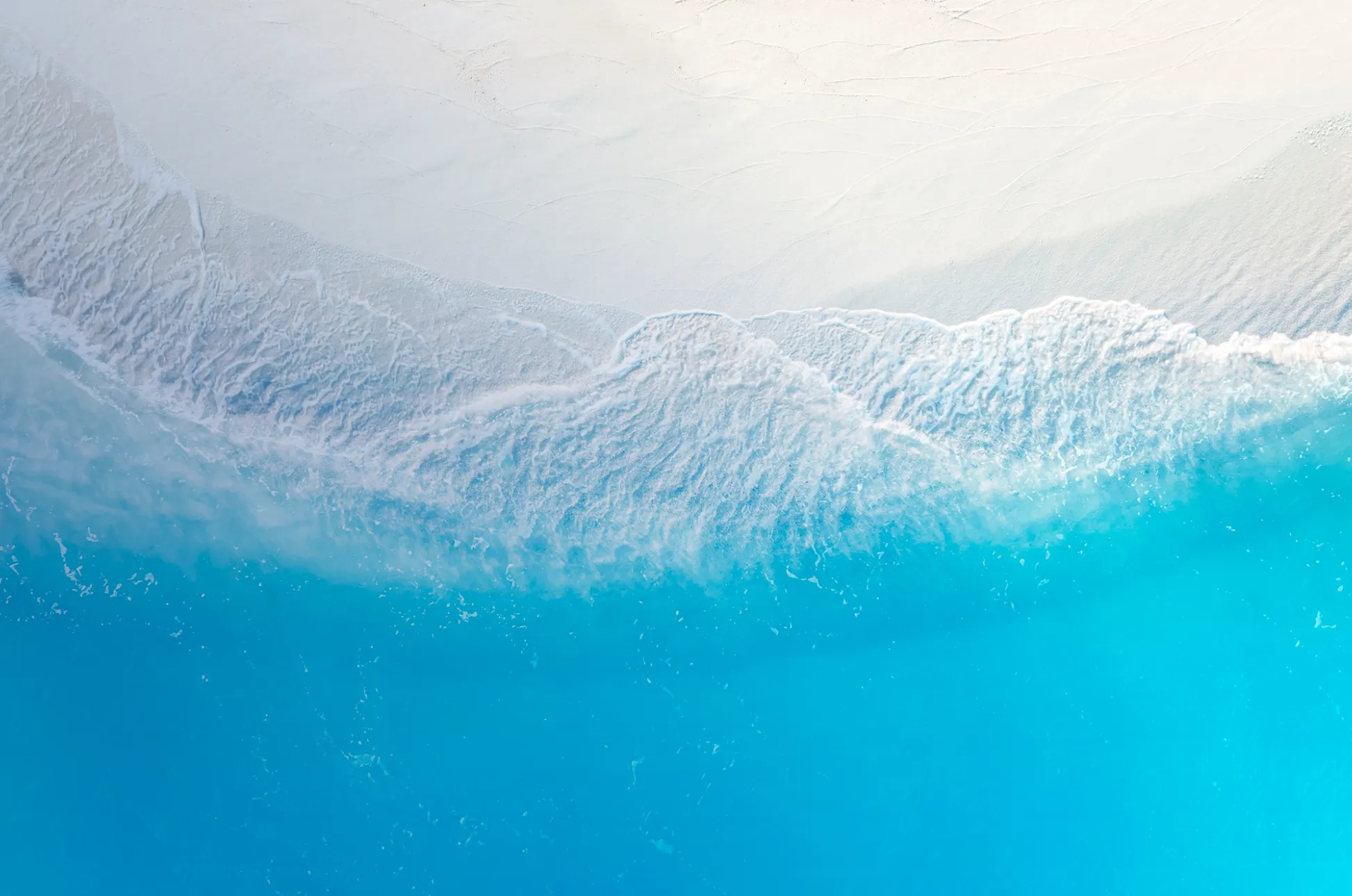 Aerial view of gentle ocean waves washing onto a light sandy beach with clear blue water.