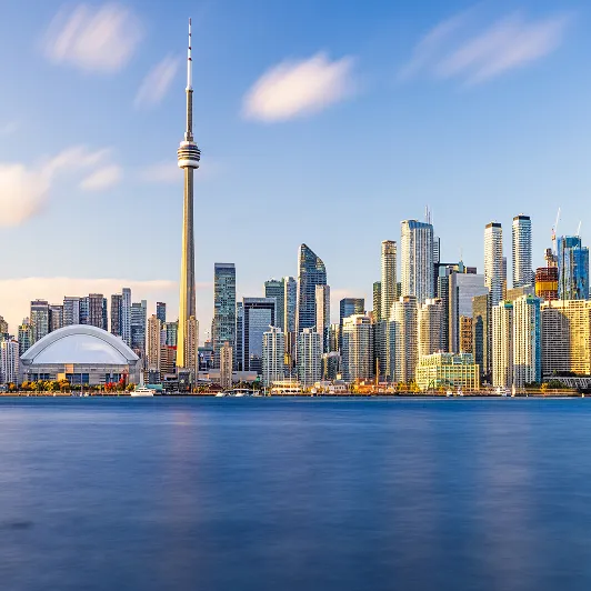 Toronto skyline featuring the CN Tower and Rogers Centre under a blue sky with clouds, viewed across calm water.