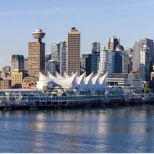 Vancouver skyline with tall buildings and Canada Place with its distinctive white sails, viewed across water.