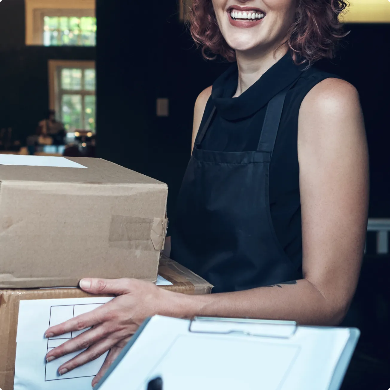 Smiling woman in black apron holding two cardboard boxes with a clipboard in front.