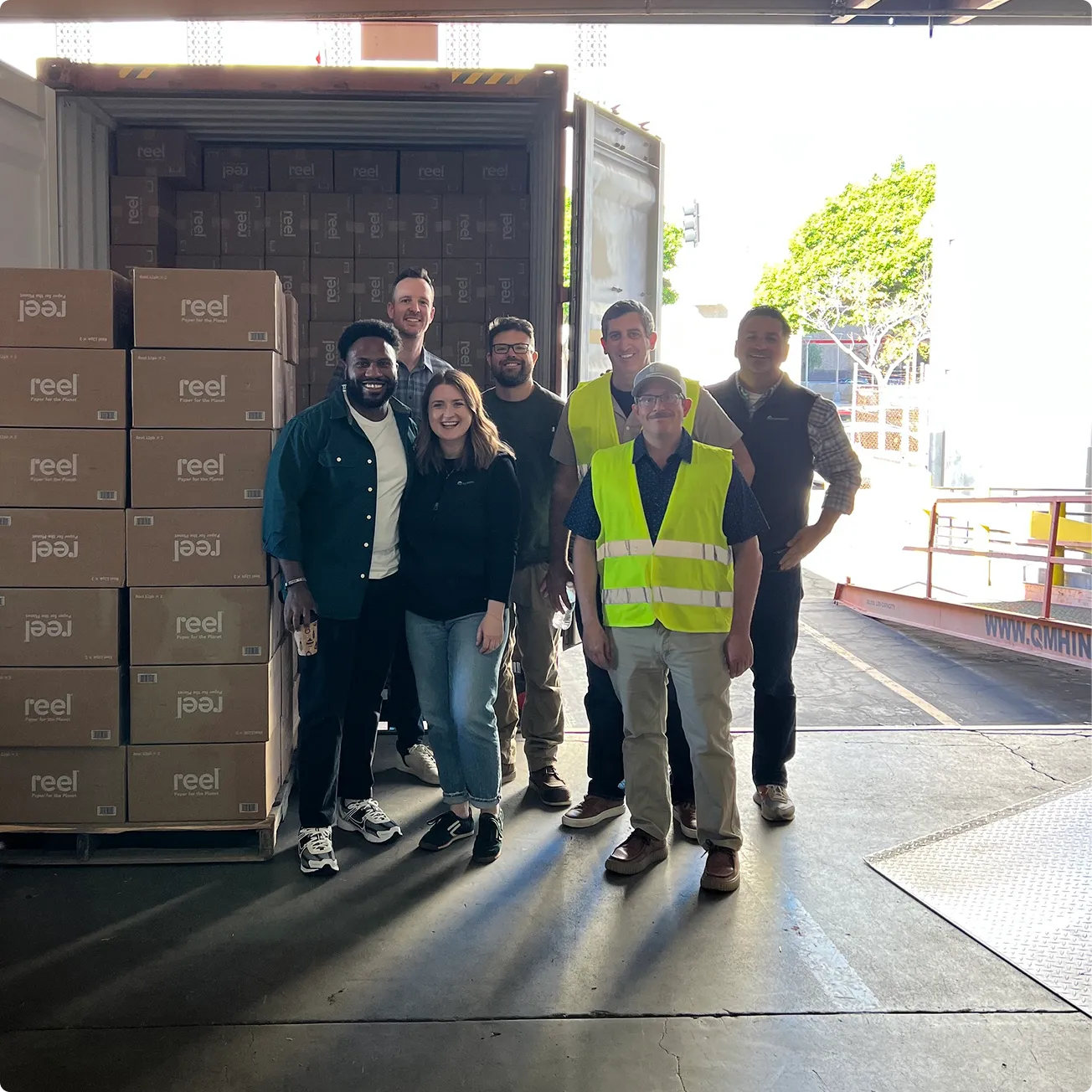 Group of seven people standing inside a warehouse near stacked boxes labeled 'reel'.