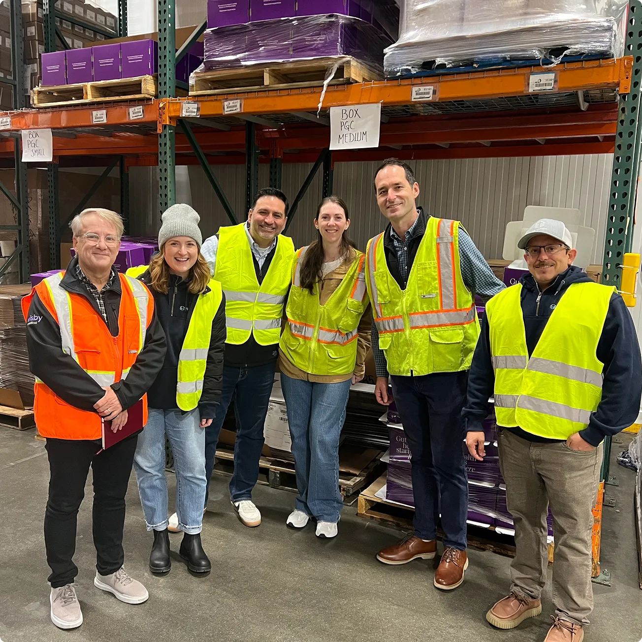 Six people wearing reflective safety vests standing and smiling inside a warehouse with shelves and boxes in the background.