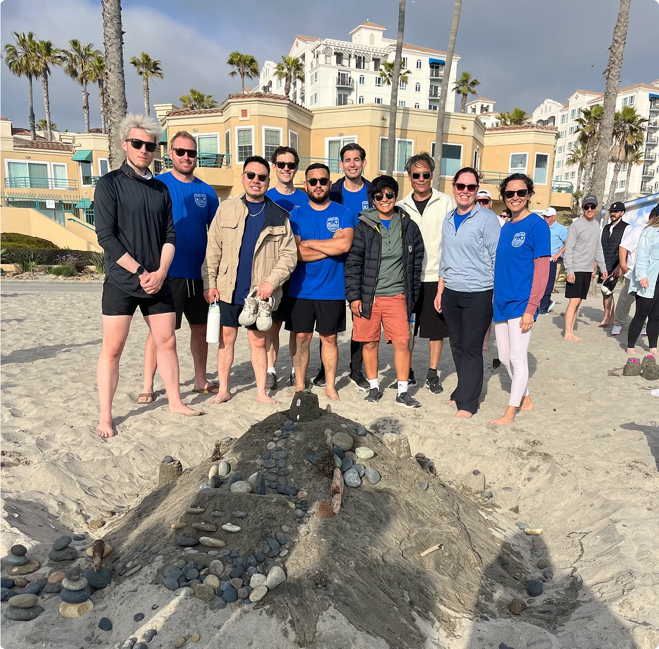 Group of eleven people standing on a beach behind a large decorated sandcastle, with buildings and palm trees in the background.
