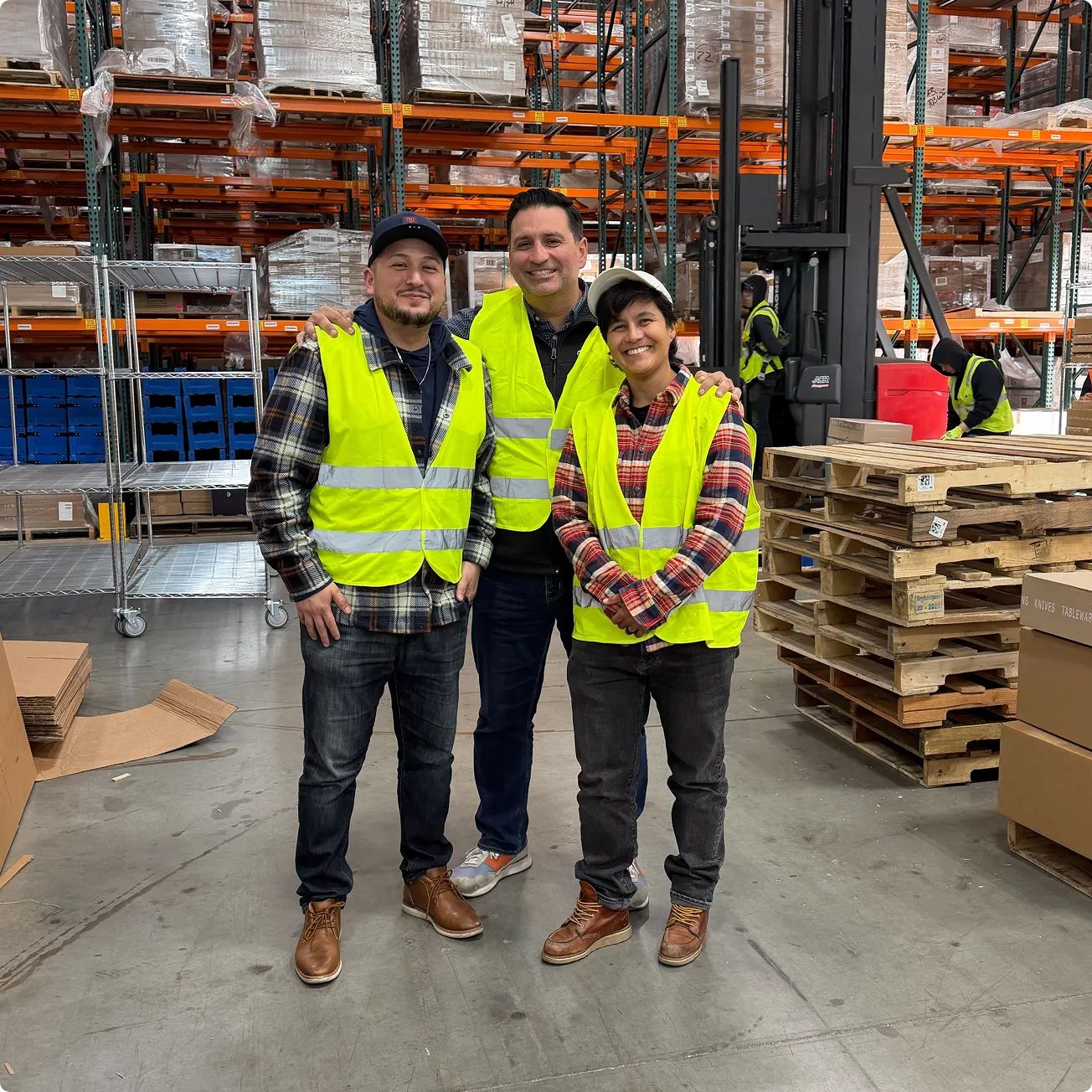 Three warehouse workers wearing yellow safety vests posing and smiling inside a storage facility with pallets and shelves.