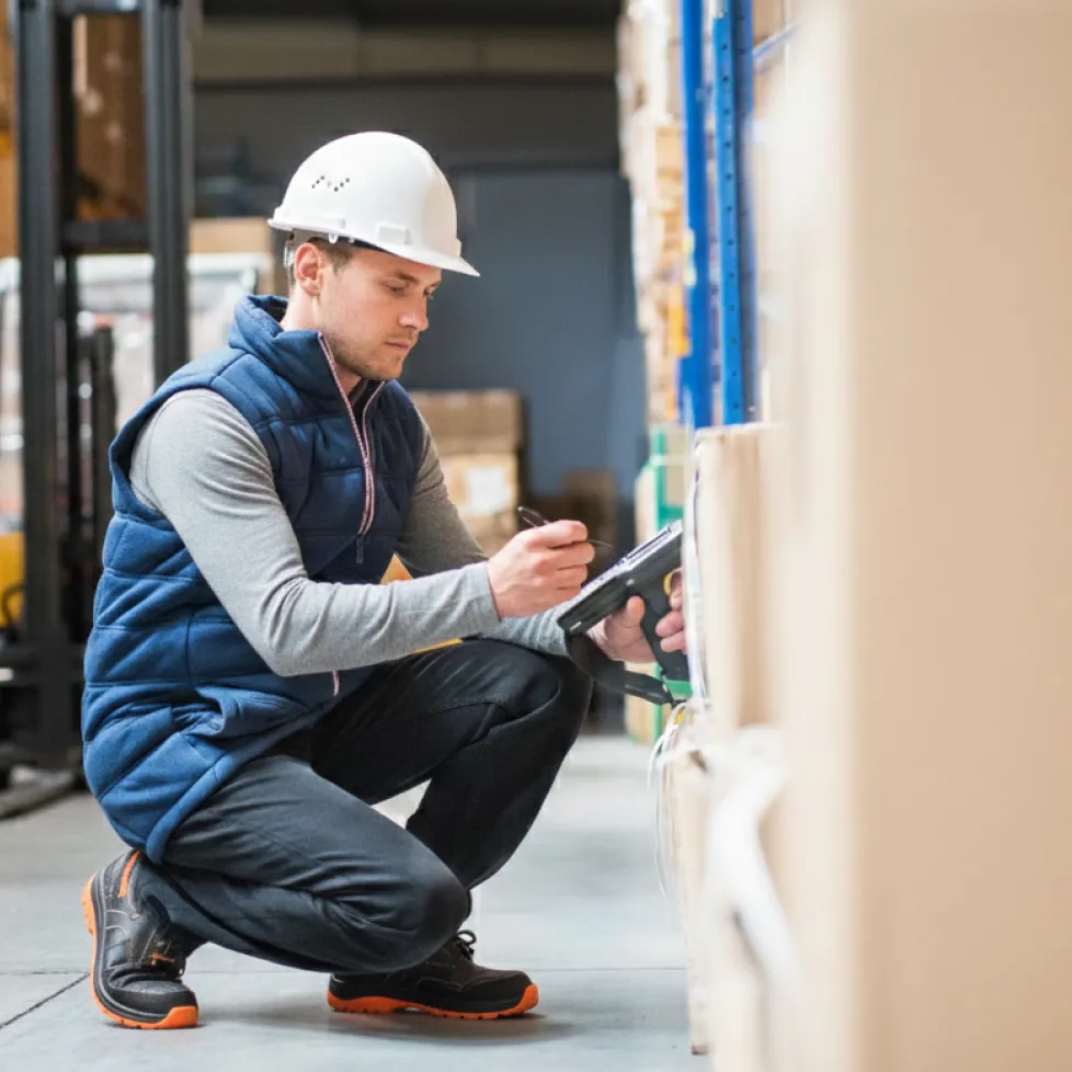 Warehouse worker in a white hard hat and blue vest scans and checks inventory on stacked boxes.
