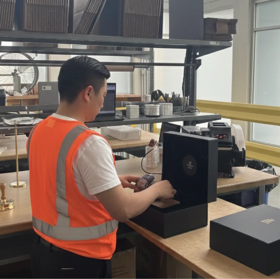 Man in orange safety vest unpacking a black luxury watch box at a workspace with shelves and a laptop.