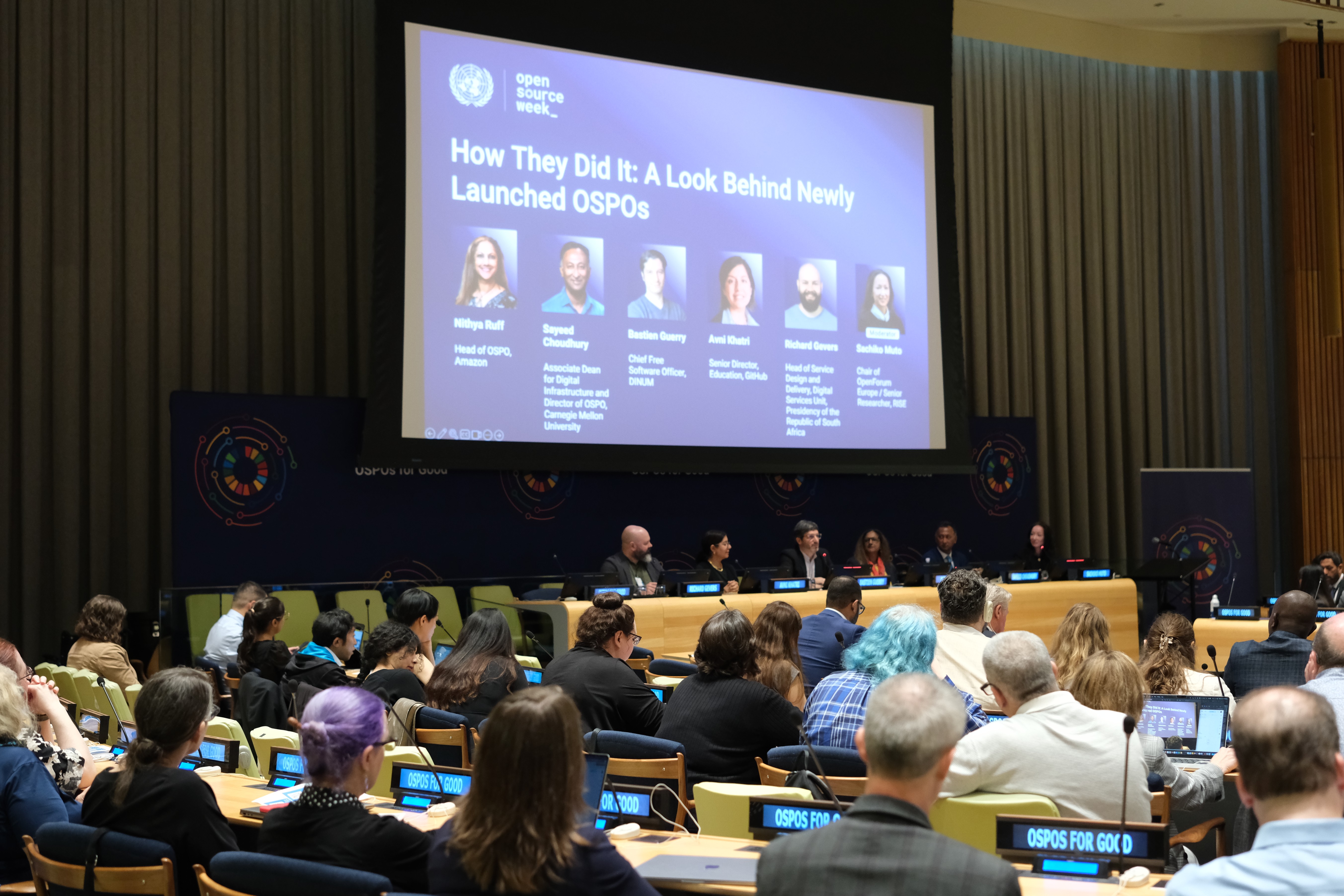 Conference panel discussing 'How They Did It: A Look Behind Newly Launched OSPOs' with multiple people seated at tables and a large screen displaying speaker names and titles.