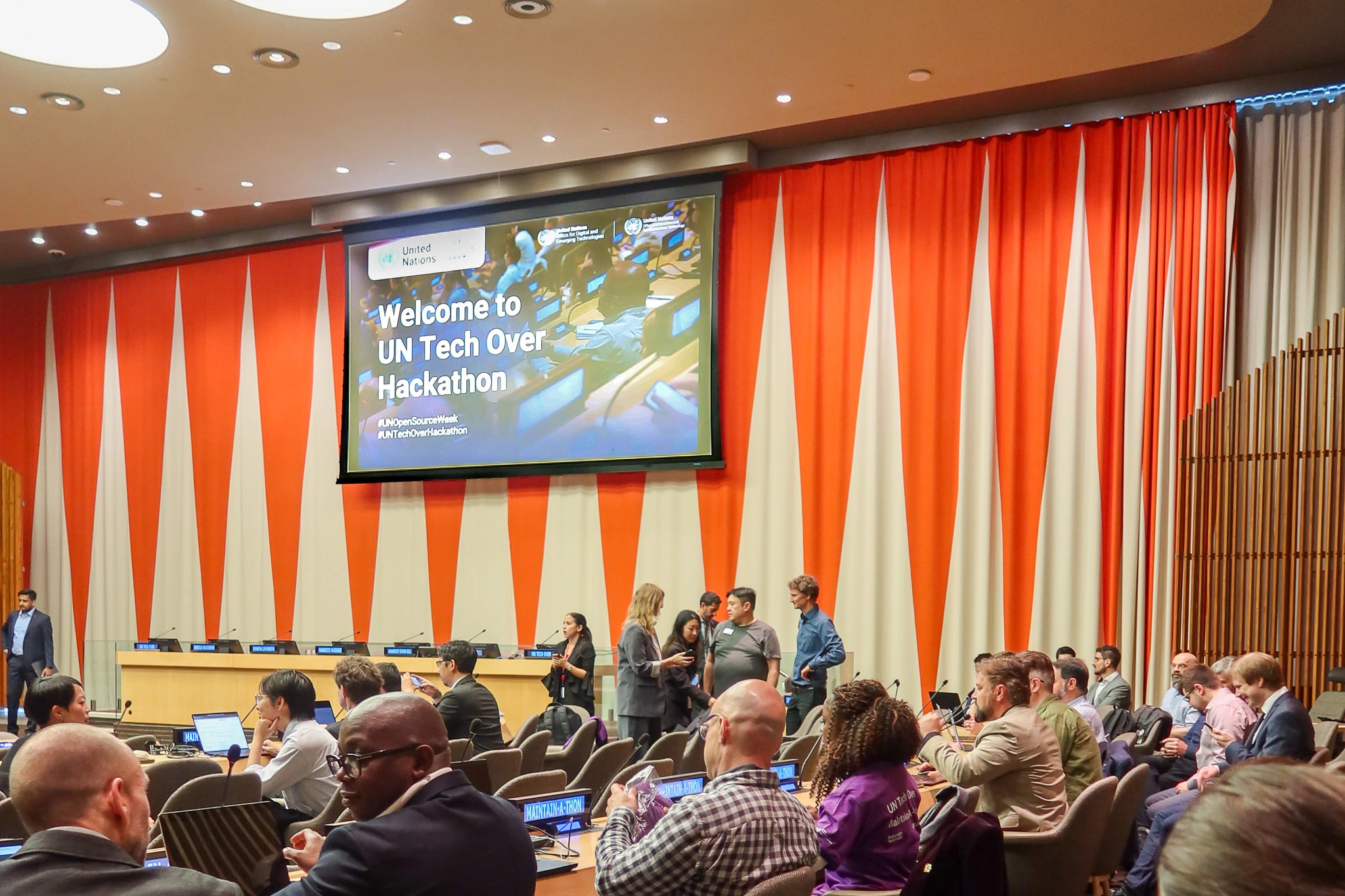 un open source week 2025, Attendees conversing and working on laptops in a conference room with orange and beige curtains, beneath a screen displaying 'Welcome to UN Tech Over Hackathon.'