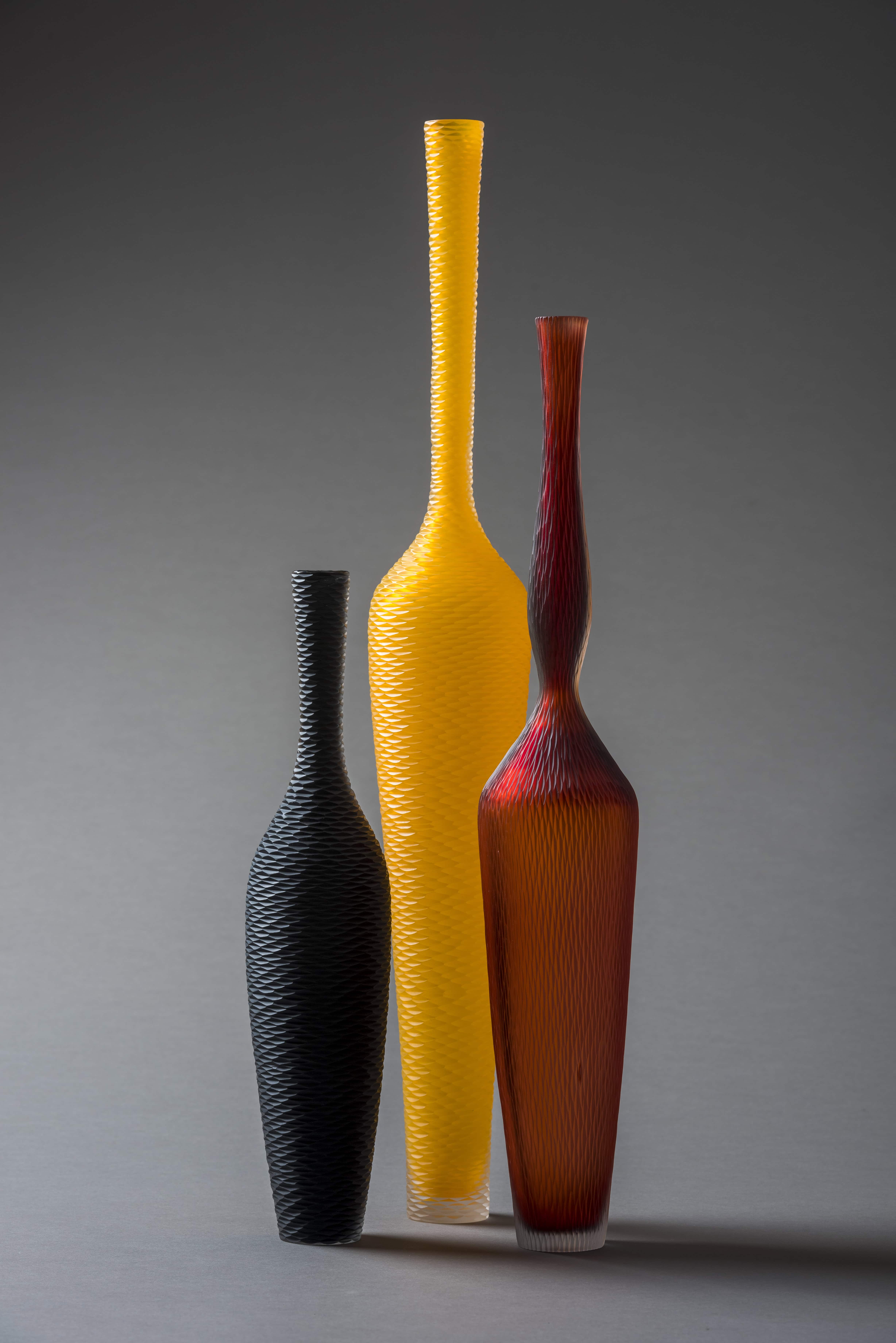 Trois vases en verre texturé aux formes allongées et couleurs noir, jaune et rouge posés sur fond gris.