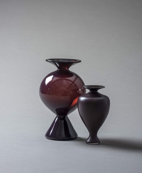 Deux vases en verre violet foncé, l'un rond avec un socle, l'autre élancé, sur fond gris.