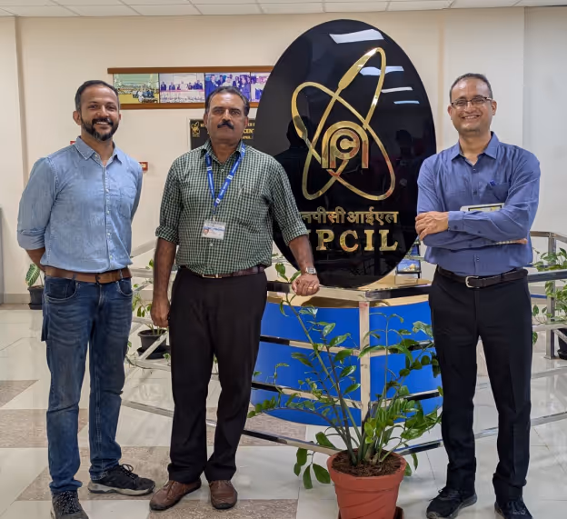 Three men standing indoors in front of a black and gold PCIL sign with a plant in a red pot nearby.