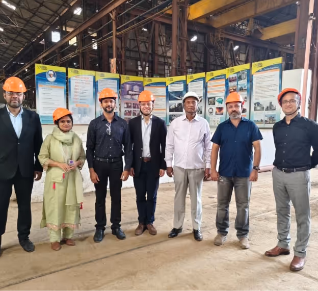 Seven people wearing safety helmets standing indoors in an industrial facility with informational posters in the background.