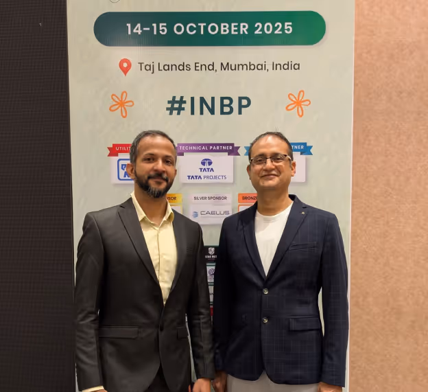 Two men in suits standing and smiling in front of a banner displaying event details for 14-15 October 2025 at Taj Lands End, Mumbai, India, with the hashtag #INBP.