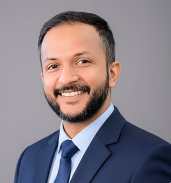 Smiling man with beard wearing a dark blue suit and tie against a plain gray background.