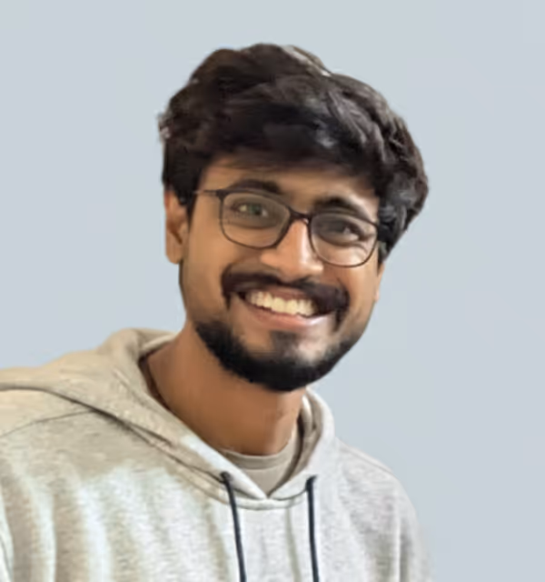 Smiling young man with glasses and a beard wearing a light gray hoodie against a plain background.