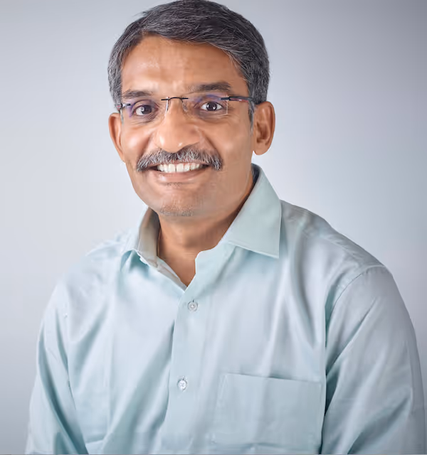 Middle-aged man with short graying hair, glasses, and a mustache, wearing a light blue collared shirt, smiling against a plain light background.