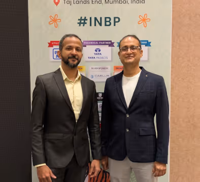 Two men in business attire standing in front of a banner with #INBP and sponsor logos at Taj Lands End, Mumbai.