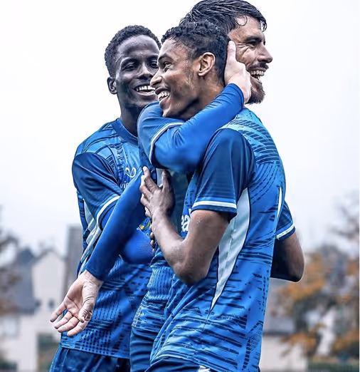 Three male soccer players in blue jerseys celebrating and smiling together outdoors.