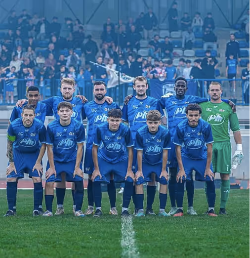 Soccer team in blue uniforms posing on the field with goalkeeper in green kit, spectators in stadium behind them.