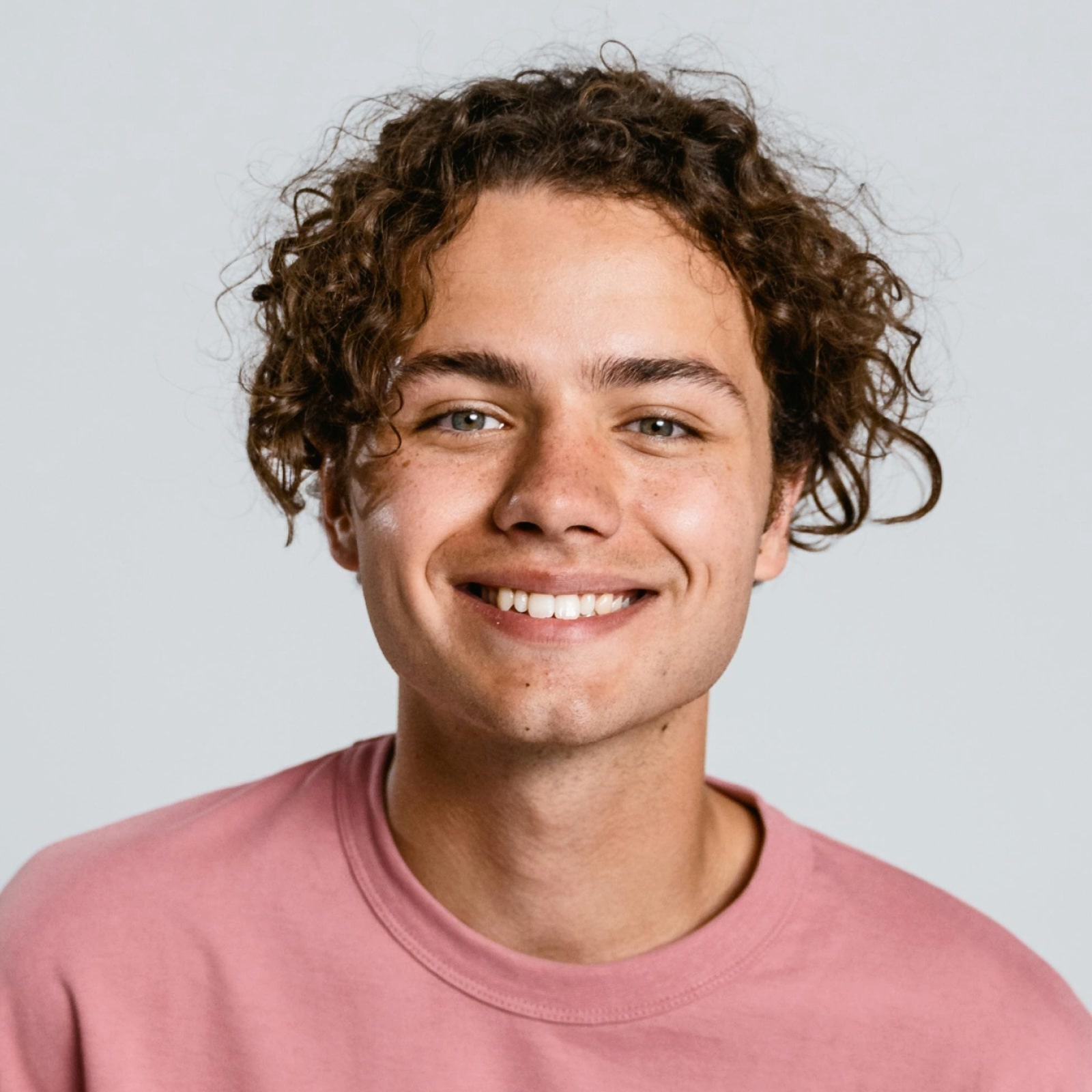 Smiling young man with curly brown hair wearing a pink sweatshirt, against a plain light background.