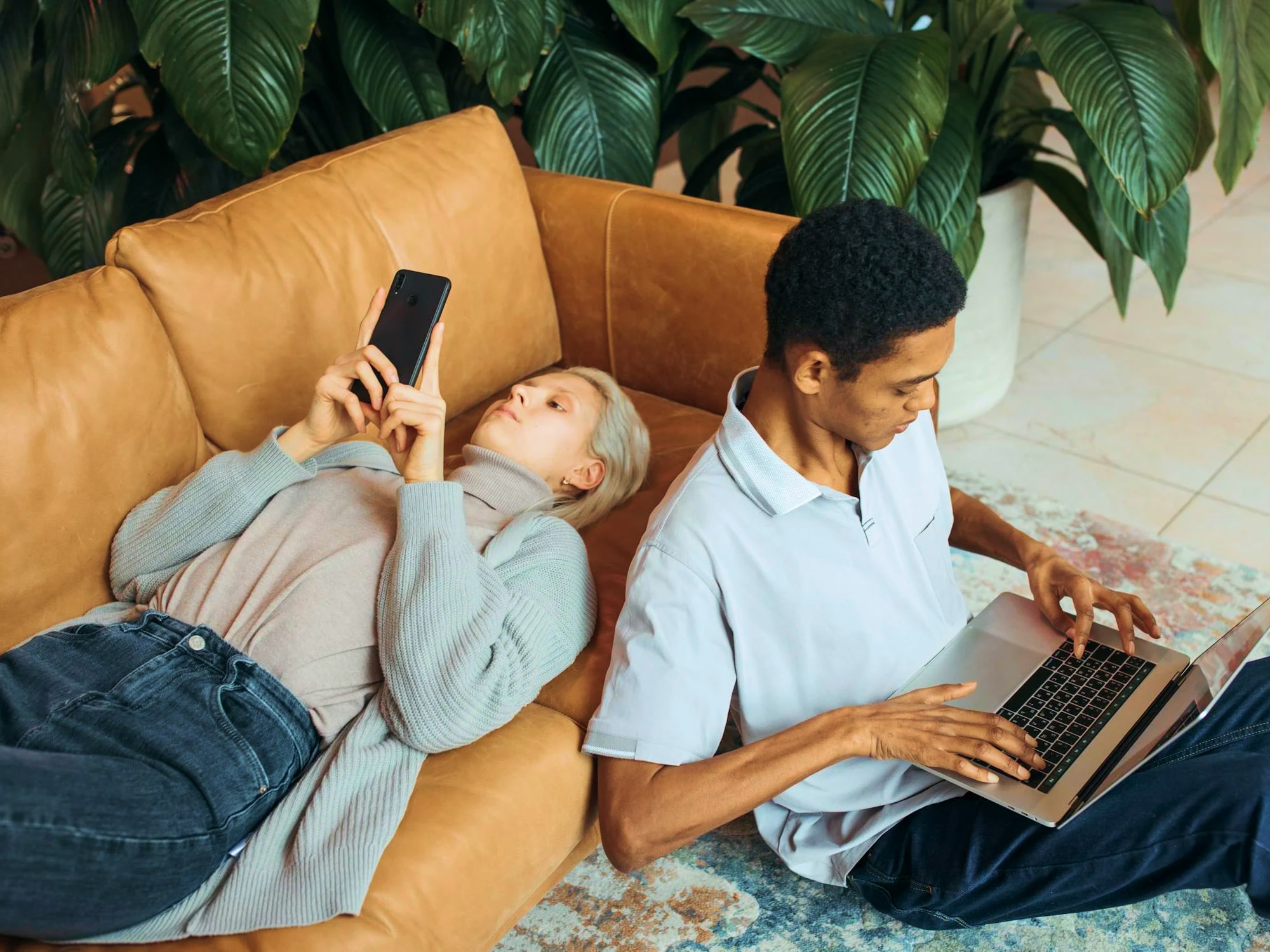 A young woman lying on a tan sofa using a smartphone and a young man sitting on the floor using a laptop, with large green plants in the background.