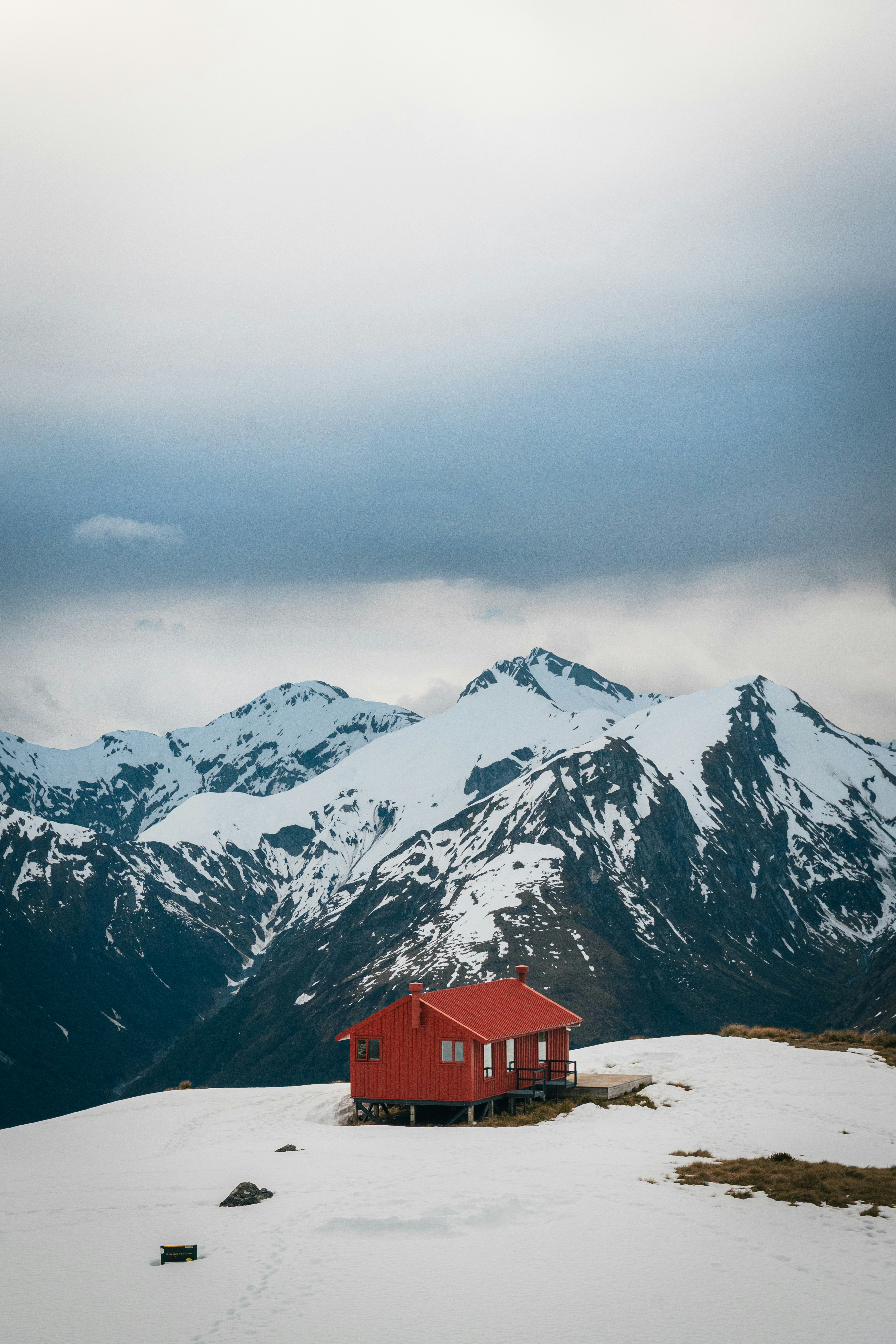 Small red cabin on snowy mountain slope with snow-covered peaks under a cloudy sky.