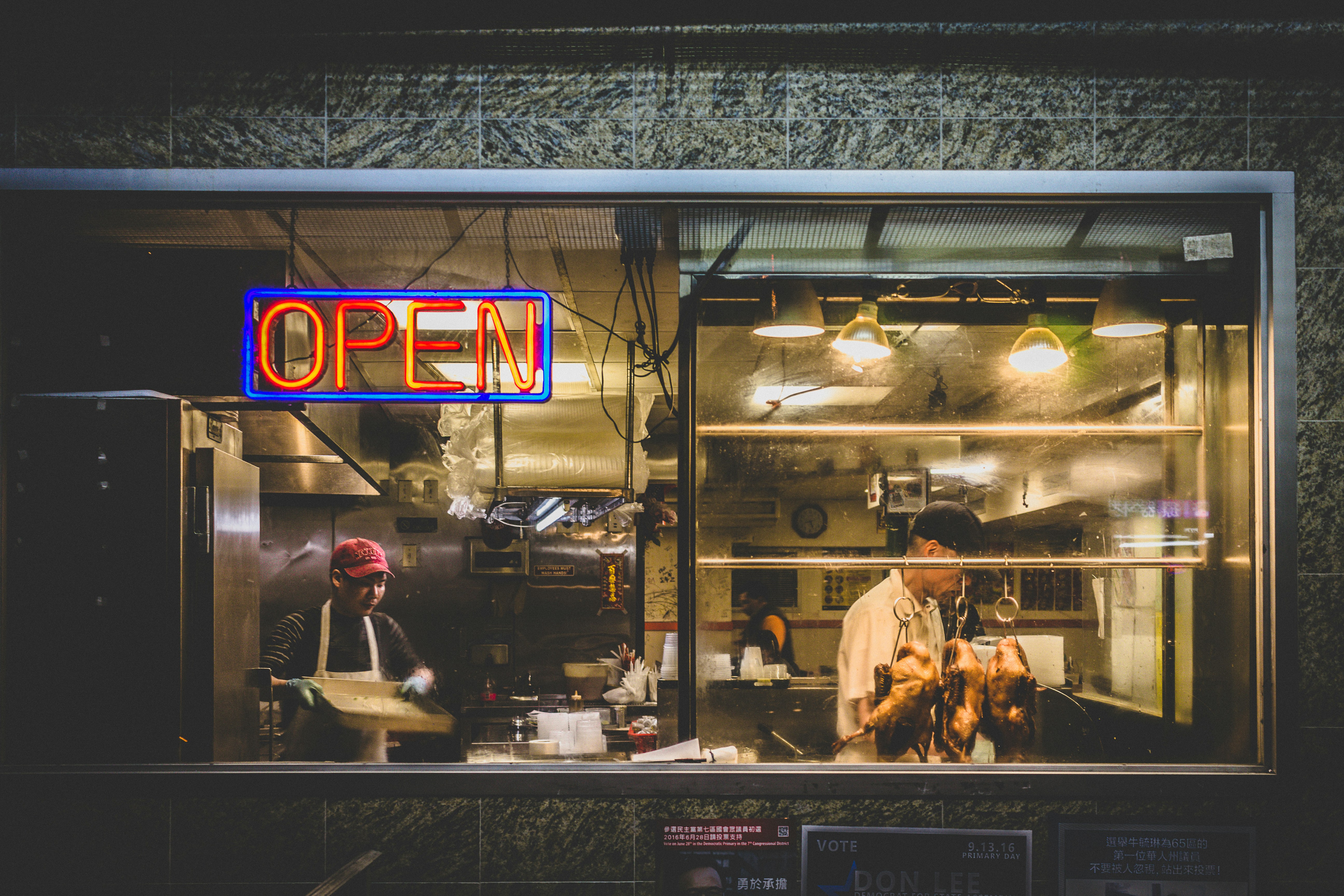 Restaurant kitchen seen through a window with an illuminated OPEN neon sign and roasted ducks hanging inside.