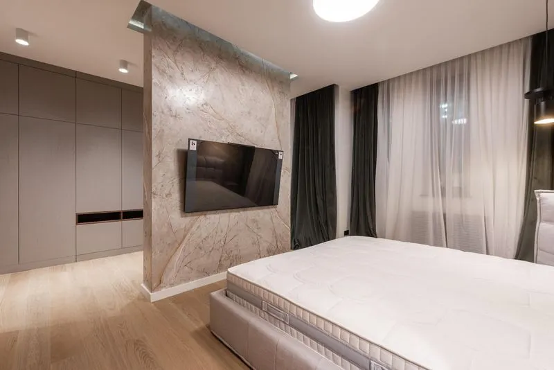 Modern bedroom with a large white mattress, marble accent wall with mounted flat-screen TV, wooden floor, and floor-to-ceiling dark and sheer curtains.
