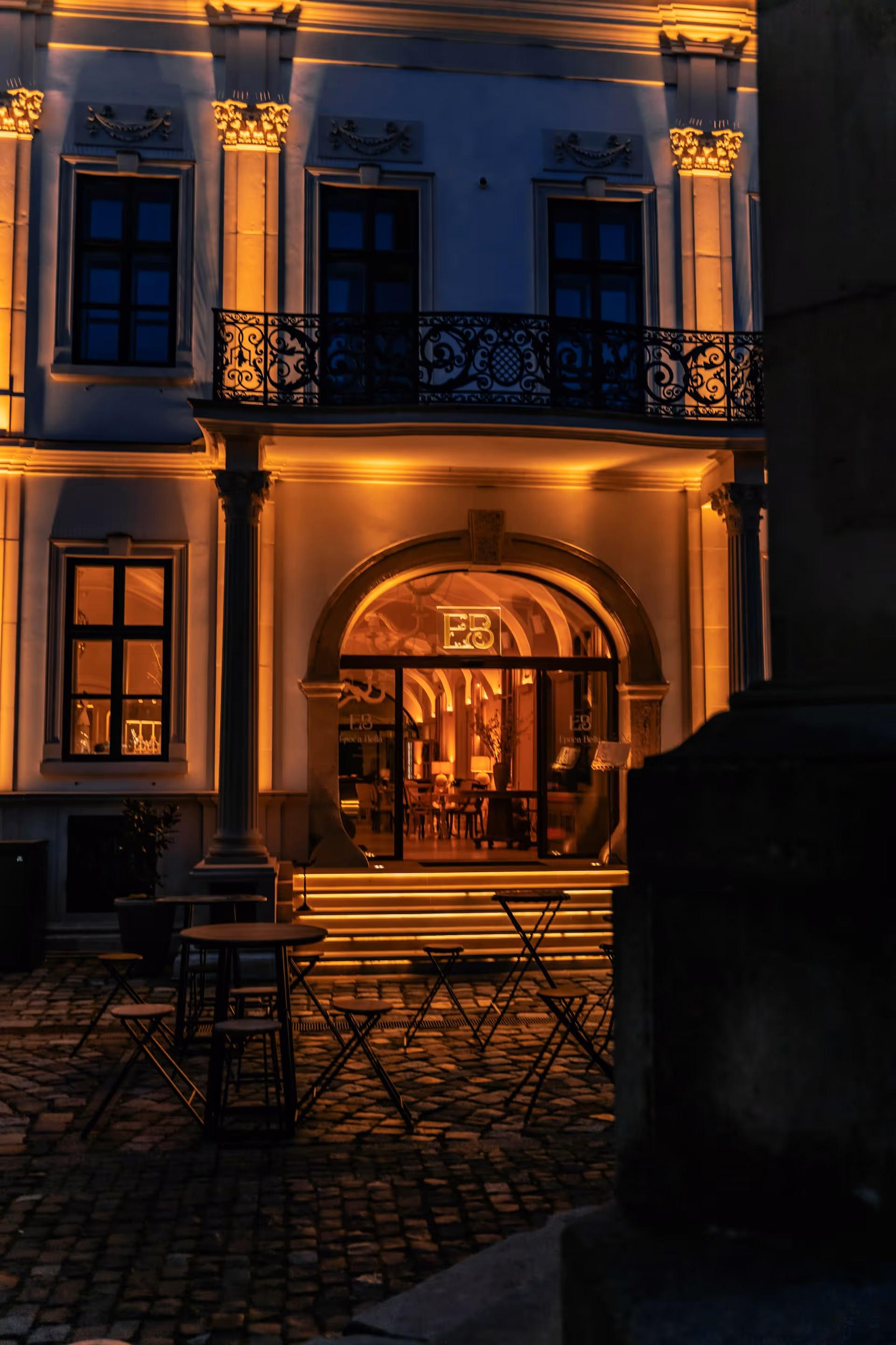Outdoor seating area with tables and chairs on cobblestone in front of a warmly lit, classical building entrance at night.