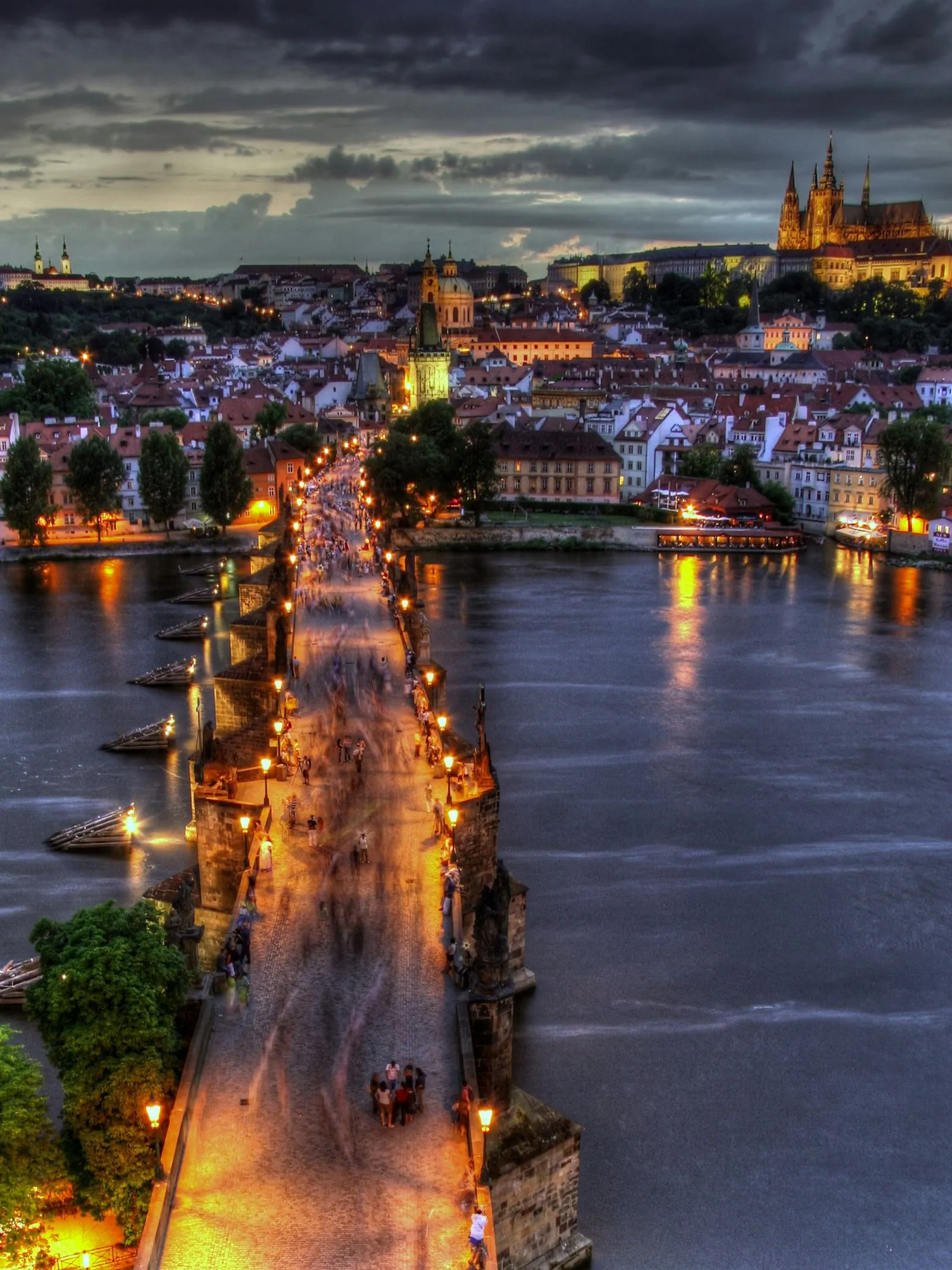 Illuminated Charles Bridge with people walking over the Vltava River at dusk in Prague, with Prague Castle visible in the background under a cloudy sky.