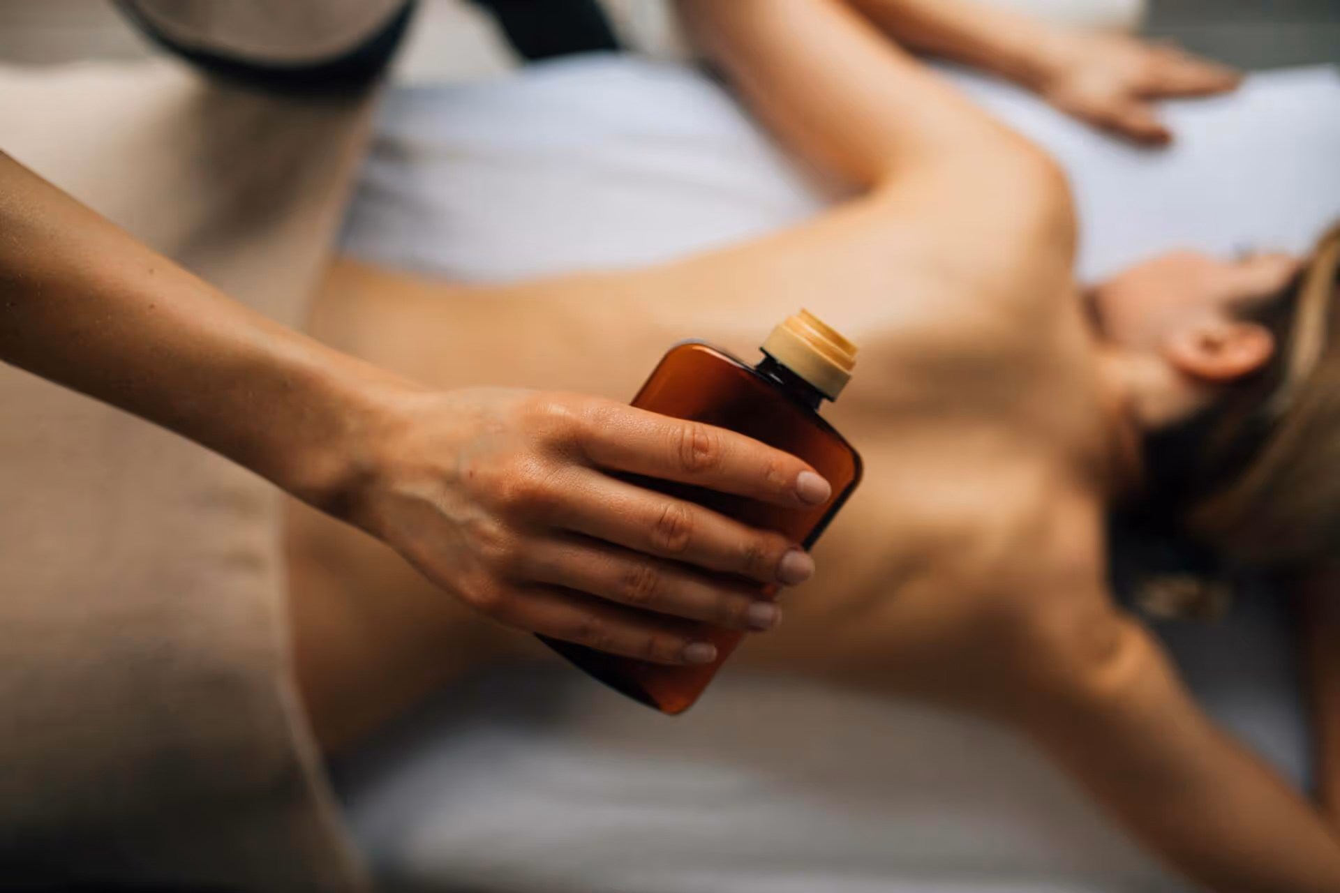 Hand holding a brown bottle of massage oil over a woman lying face down on a massage table.