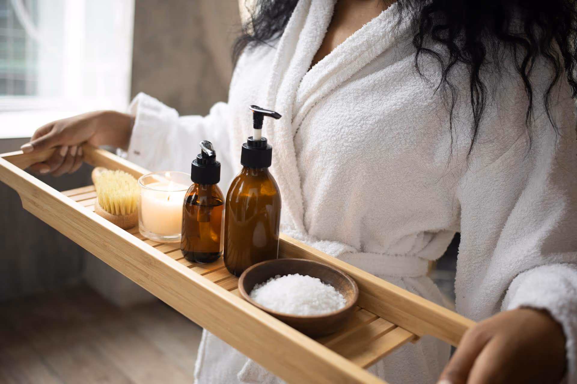 Person in white robe holding a wooden tray with two amber pump bottles, a lit candle, a brush, and a bowl of bath salts.