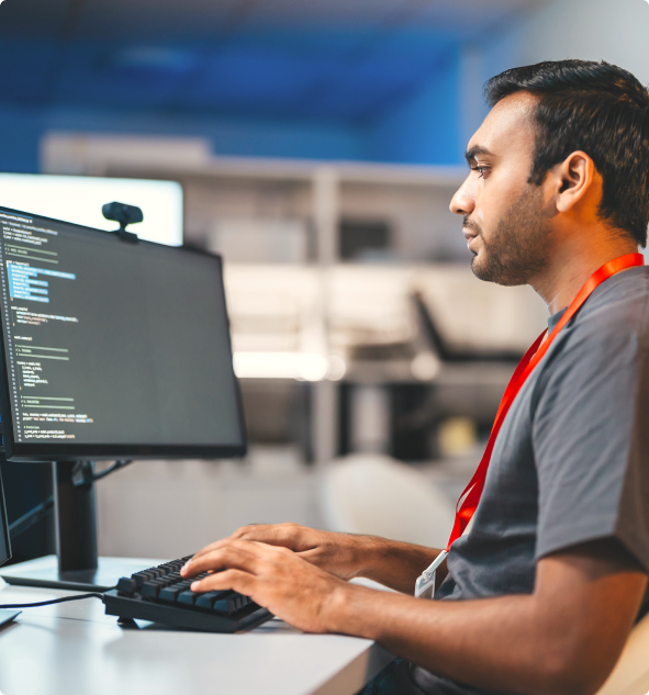 Man wearing a red lanyard typing on a keyboard while looking at a computer screen displaying code in an office setting.