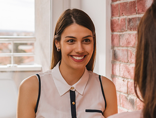 Smiling woman in a sleeveless white blouse sitting indoors near a brick wall, facing another person.