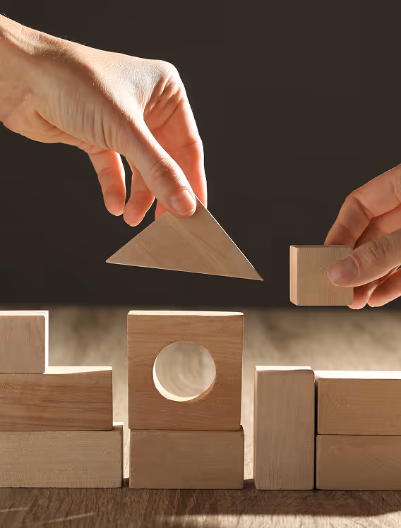 Two hands arranging light wooden blocks, including a triangle and a square block, on a wooden surface with a dark background.