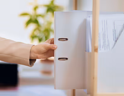 Hand placing a white lever arch file on a shelf with blurred green plant in the background.