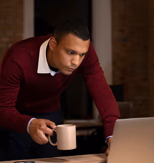 Man in a maroon sweater leaning forward while holding a coffee mug and looking at a laptop screen.