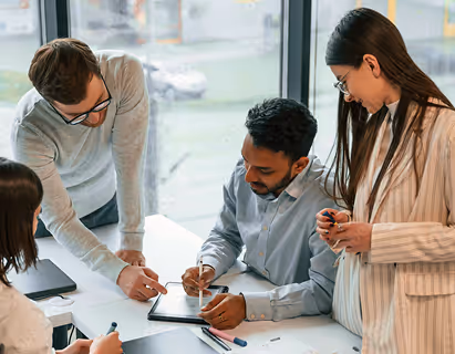 Three colleagues collaborate over a tablet in a bright office with large windows.