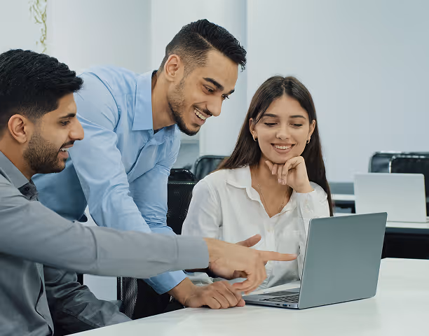 Three young professionals smiling and discussing work around a laptop in a bright office.