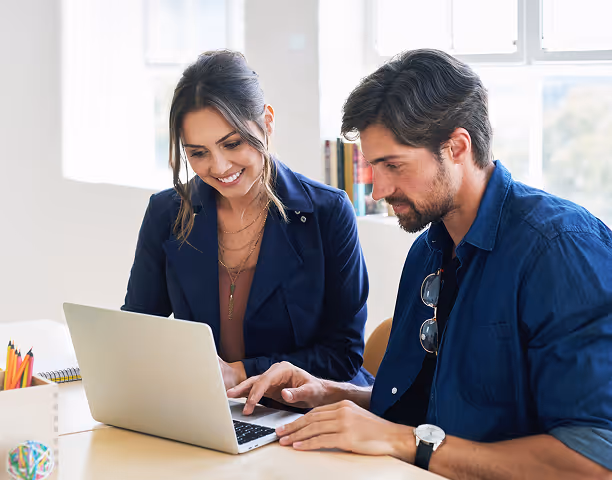 A man and woman collaborating while looking at a laptop on a desk in a bright office.